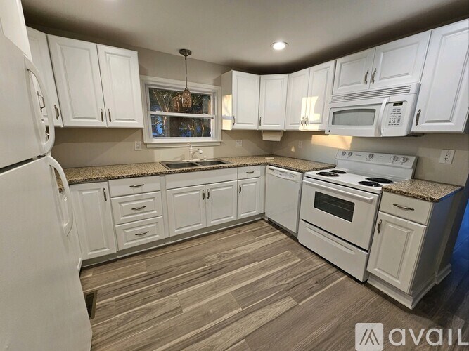 A kitchen with white cabinets and appliances, a wooden floor, and a window with a view of trees.