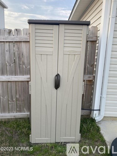 A small, light-colored wooden shed with a black handle is situated in a backyard.