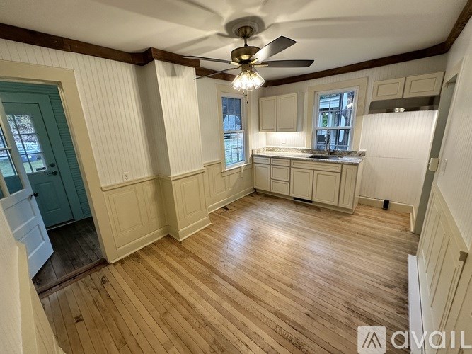A room with wooden floors and white walls, featuring a kitchen area with cabinets and a window.