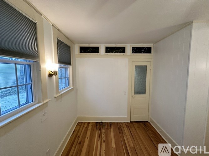 A room with wooden floors and white walls, with a window on the left and a door on the right.