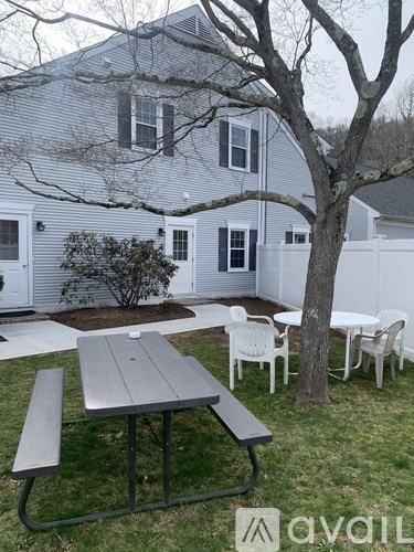 A grey picnic table is in the foreground of a yard with a white fence and house in the background.