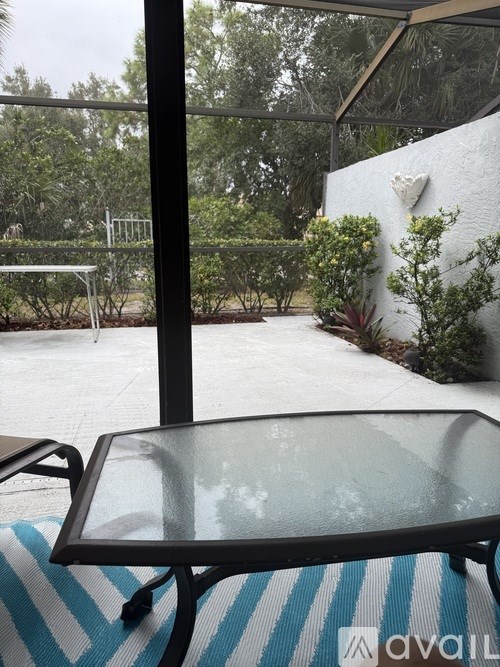 A table with a glass top and metal frame is on a striped rug in front of a patio with a white wall and greenery.
