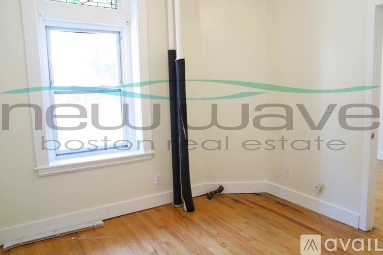 A room with a wooden floor and a window, with a large black object leaning against the wall.