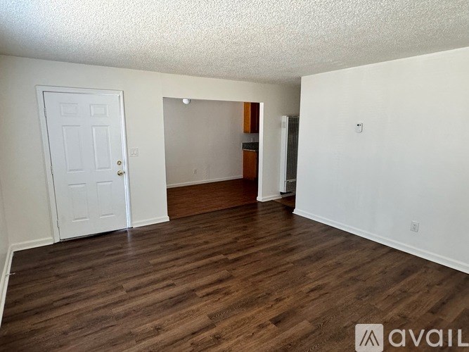 A room with wooden flooring and white walls, with a door on the left and a partially visible doorway on the right.