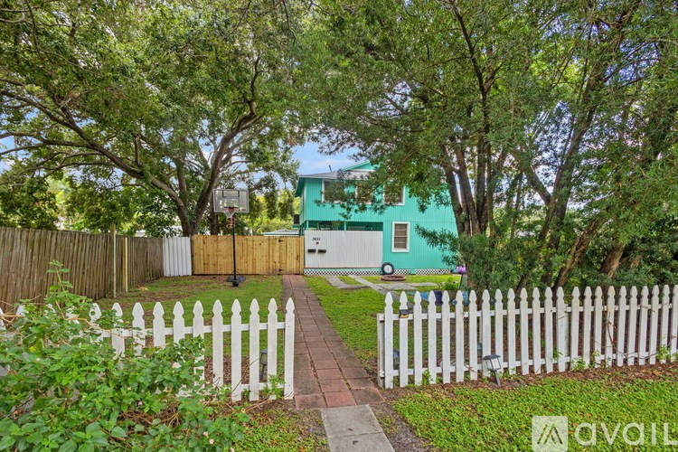 A green house is surrounded by a white picket fence and trees.