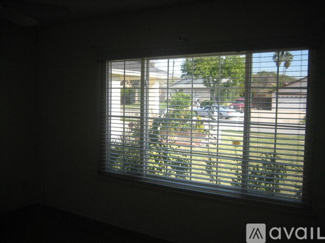 A window with blinds is open, letting in light and showing a view of a street with cars and houses.