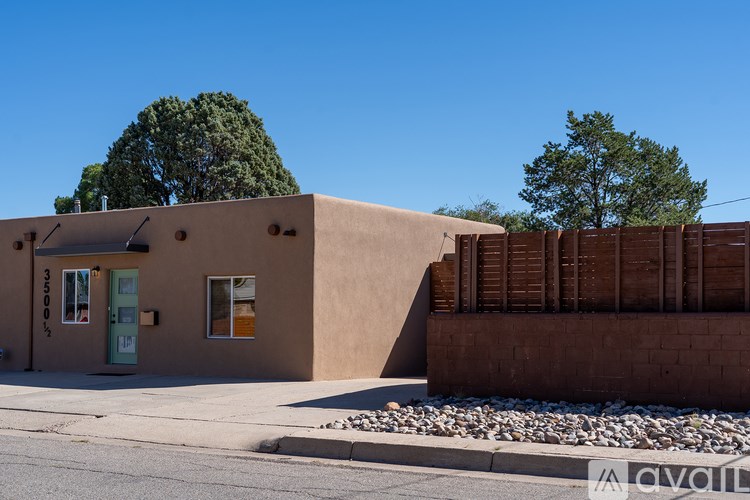 A small building with a brown facade and a green door is surrounded by a wooden fence and a tree.