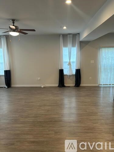 A spacious room with a ceiling fan and lighting, featuring a large window with curtains and a wooden floor.