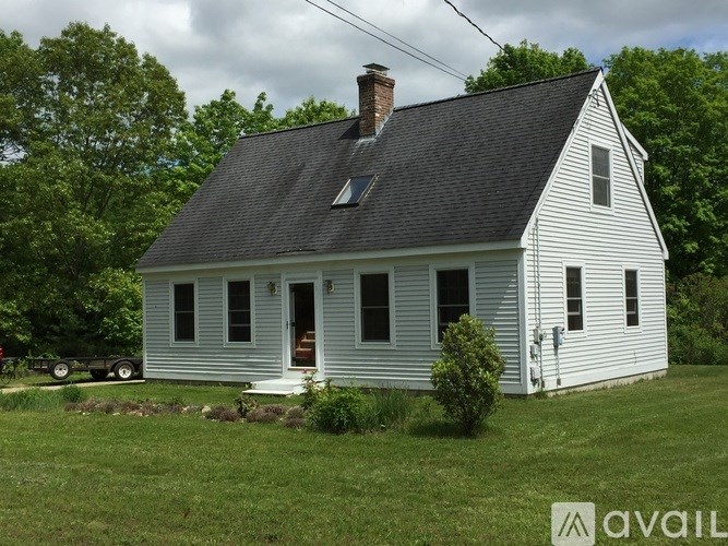 A white house with a black roof and a chimney is surrounded by greenery.