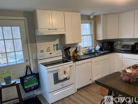 A kitchen with white appliances and cabinets, a bag on the floor, and a table with a bowl of fruit on it.