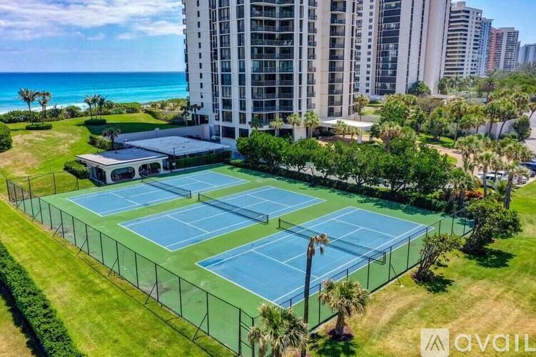 A tennis court surrounded by a fence and trees with a building in the background.
