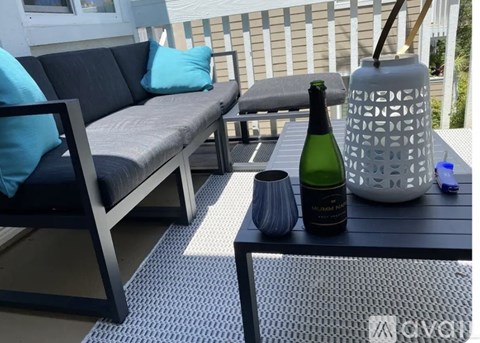 A bottle of champagne sits on a table with a grey couch and a white vase in the background.