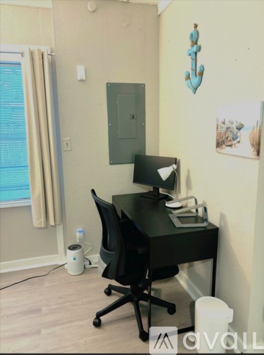 A black computer desk with a monitor, keyboard, and mouse is set up in a room with a window and a picture on the wall.