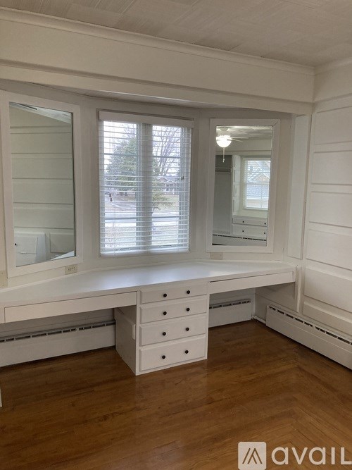 A white desk with drawers is in a room with wooden floors and a window with blinds.