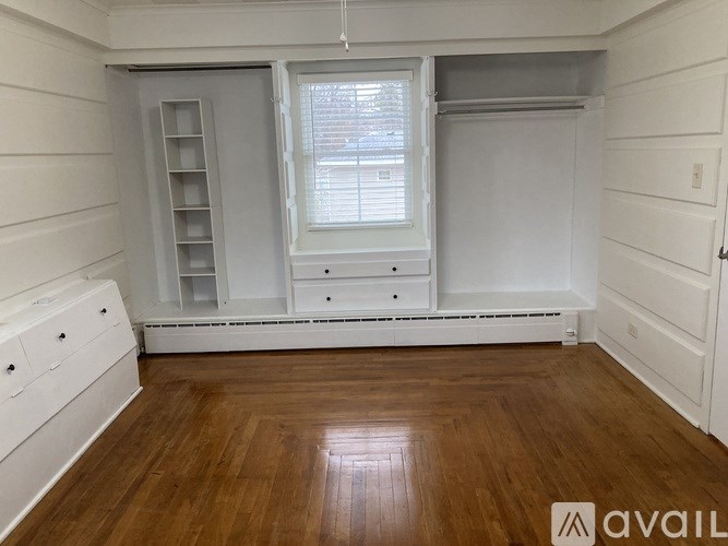 A room with wooden floors and white walls, featuring a window with blinds, a built-in shelf, and a white drawer unit.