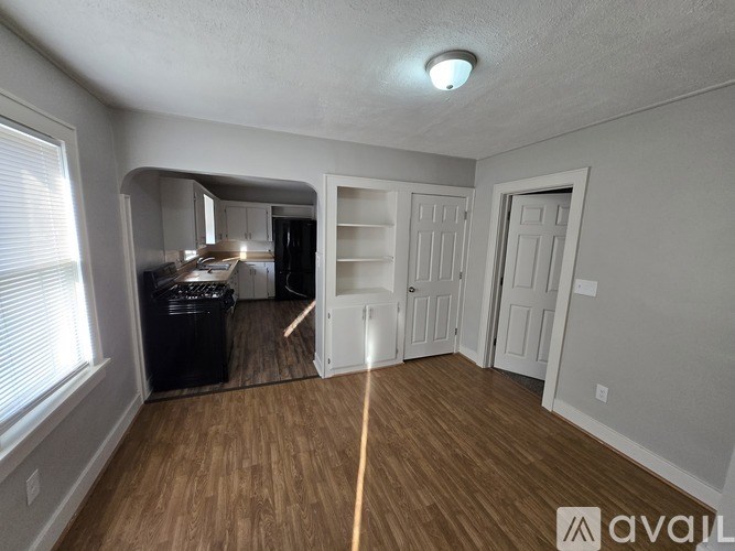 A room with a wooden floor and white walls, with a window on the left and a trash bin in the corner.