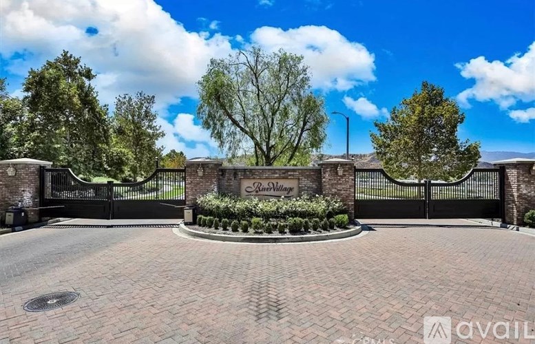 The image shows a gated entrance to a property named "Lake Village" with a circular flower bed in the foreground and trees in the background.