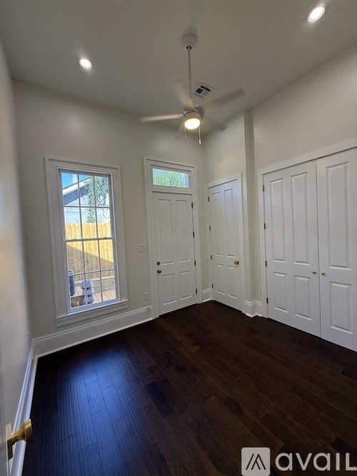 A room with white walls and wooden floors, featuring a ceiling fan and three windows.