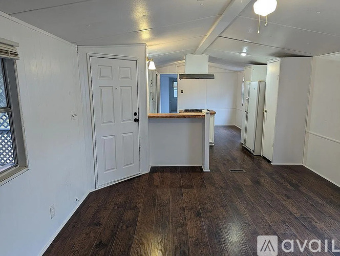 A room with wooden floors and white walls, with a door on the left and a kitchenette in the middle.
