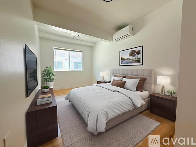 A well-lit bedroom with a large bed, a flat-screen TV, and a window with a view of a tree-lined street.