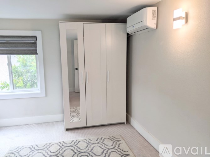 A white wardrobe with a mirrored door is in a room with a window and a wall-mounted air conditioner.