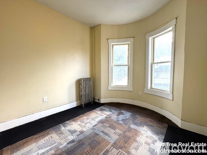 A room with a carpeted floor, two windows, and a radiator.