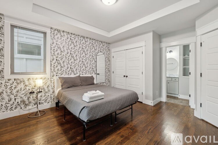 A bedroom with a patterned wallpaper, a bed with a grey bedspread and a white towel on it, a wooden floor, and a window with blinds.