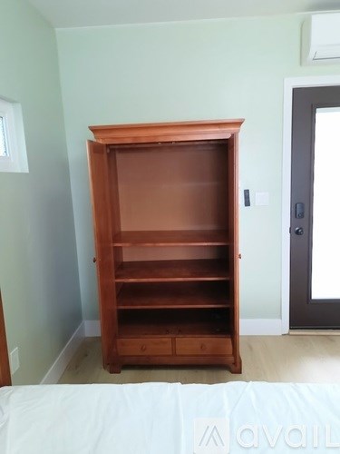 A wooden bookcase with a glass door stands in a room with a white bed in the foreground.