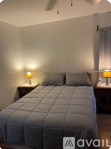 A bed with a grey comforter and two pillows sits in a room with two lamps on bedside tables.