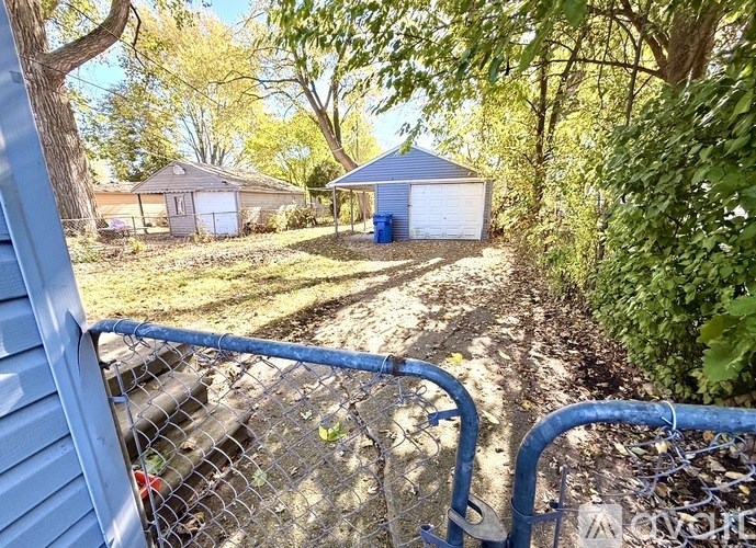A blue fence with a gate is in the foreground of a rural scene with a shed and a garage in the background.