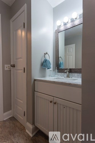 A bathroom with a white door, a mirror with lights on it, and a white cabinet with a blue towel hanging on it.