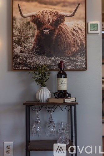 A picture of a yak is hanging on the wall above a shelf with a bottle of wine and glasses.