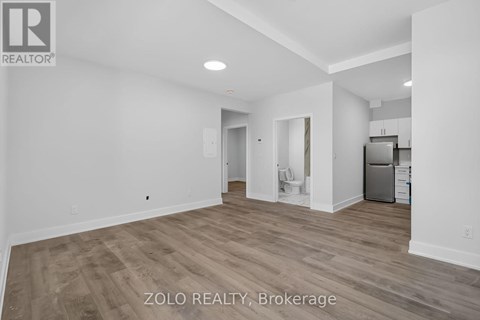 A spacious room with wood flooring and white walls, with a kitchen area visible in the background.