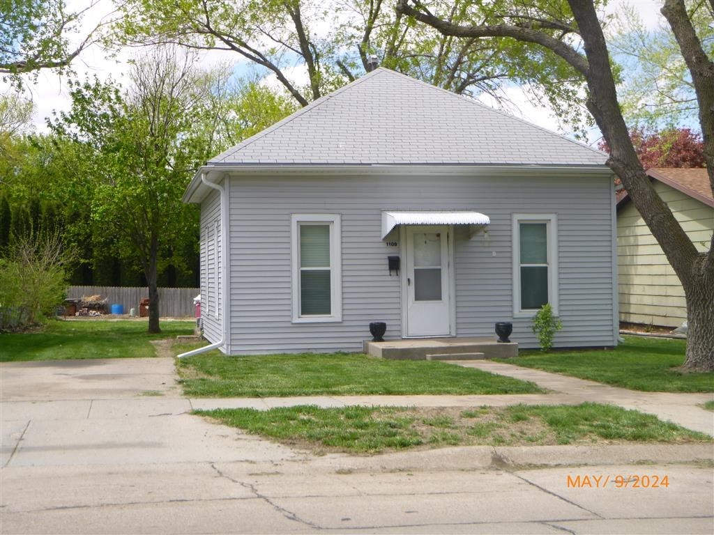 A small house with a grey roof and a white door is surrounded by greenery.