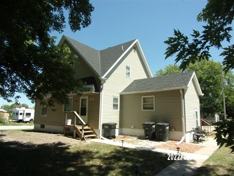 A house with a brown and beige exterior is surrounded by trees and has a driveway leading to the front door.