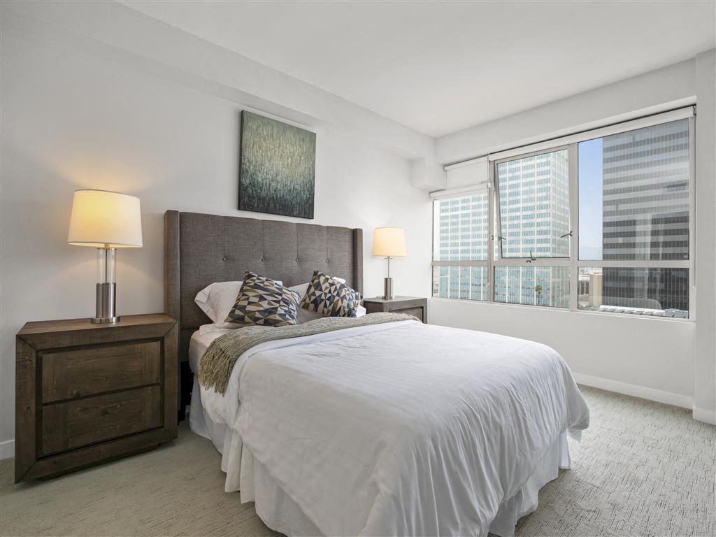 A bedroom with a large bed, a nightstand with a lamp, and a window with a view of a cityscape.