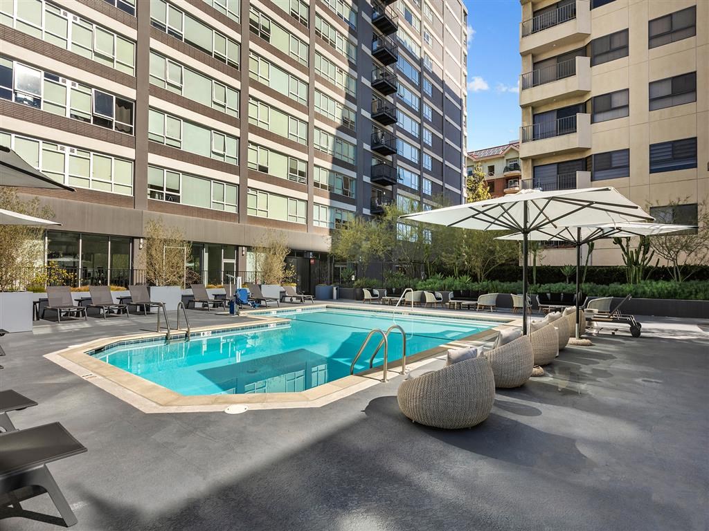 A large swimming pool surrounded by lounge chairs and umbrellas in front of apartment buildings.