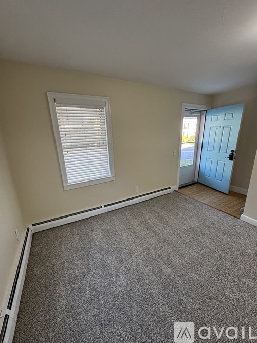 A room with a carpeted floor, a window with blinds, and a door leading to another room.