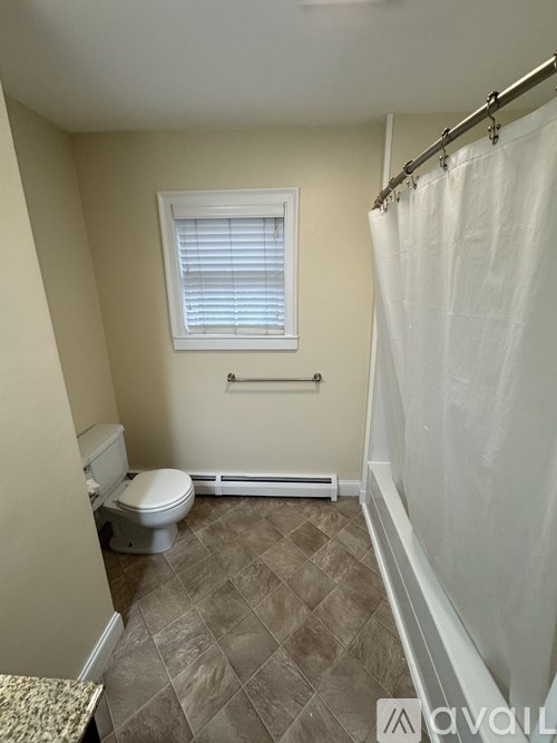 A bathroom with a toilet, a window with blinds, a tiled floor, and a shower curtain.