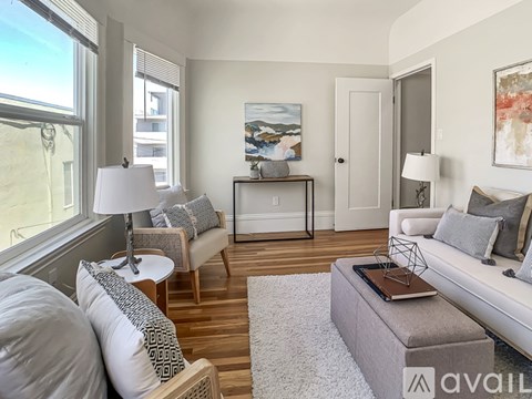 A living room with a grey couch, a white chair, a grey ottoman, a small table with a lamp, a painting on the wall, and a door.