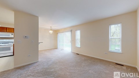 A spacious living room with beige walls and carpet flooring, featuring a kitchen area to the left and two windows with blinds.