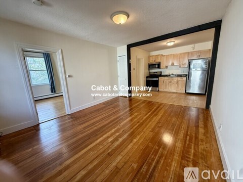 A room with wooden flooring and a kitchen area in the background.