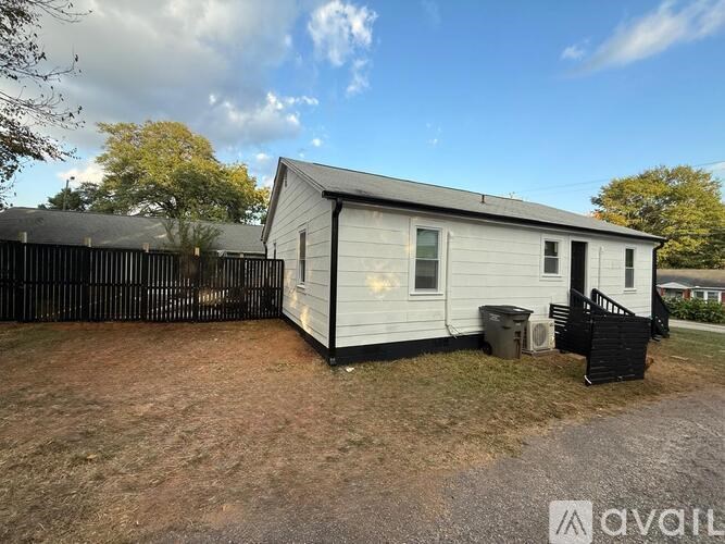 A small white house with a black fence and a black trash bin in front of it.