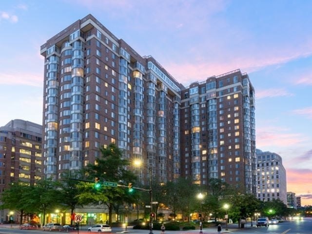 A large apartment building is lit up at dusk with cars on the street in front.