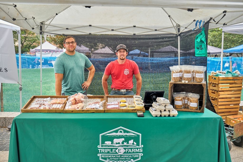 Two men are standing behind a table with a green tablecloth that has the logo of Triple C Farms on it.
