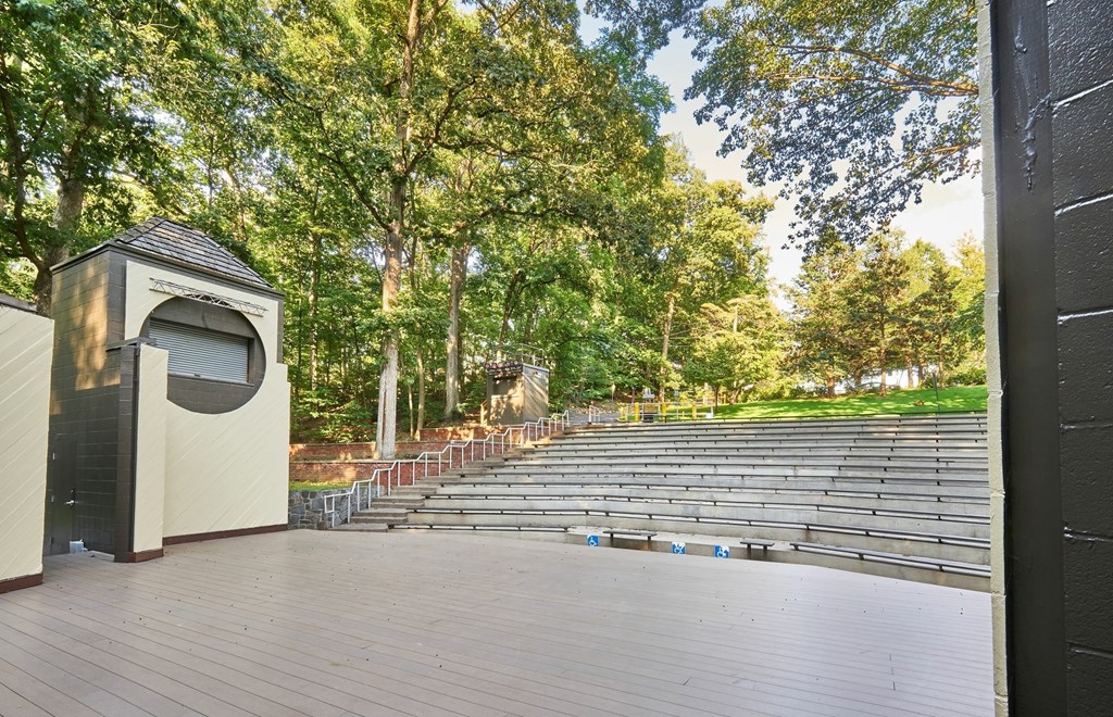 A small building with a curved window is situated in a park with a stage and seating area.