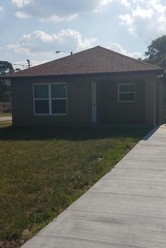 A small brown house with a white door and windows is situated on a concrete walkway.