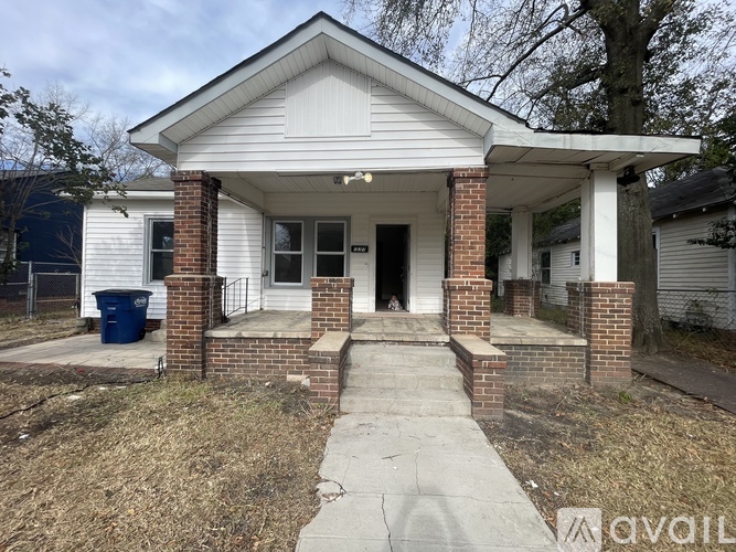 A small white house with a porch and a trash can in front.