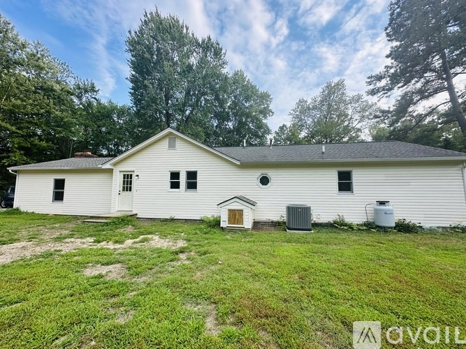 A white house with a brown roof and a brown door is surrounded by green grass and trees.