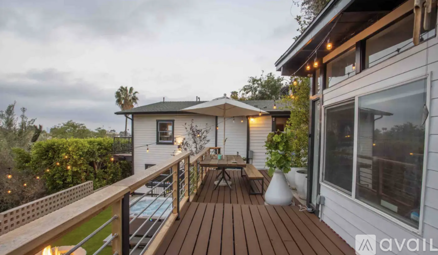 A deck with a table and chairs is surrounded by a white house and greenery.
